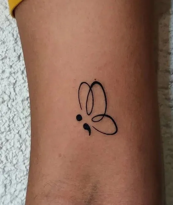 Tatuagem de ponto e vírgula onde o símbolo é acompanhado por asas de borboleta.