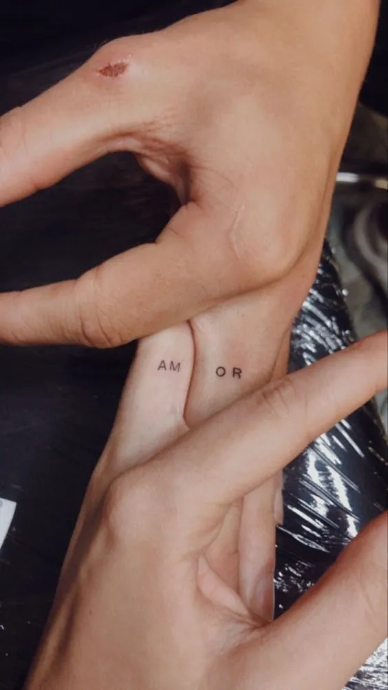 Tatuagem de casal nos dedos escrito "AM" e "OR" formando 'amor' quando os dedos se juntam